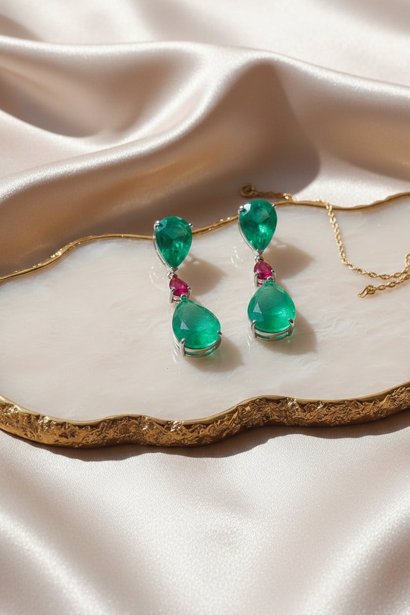 Pair of emerald green teardrop earrings with pink gemstones on a beige fabric background